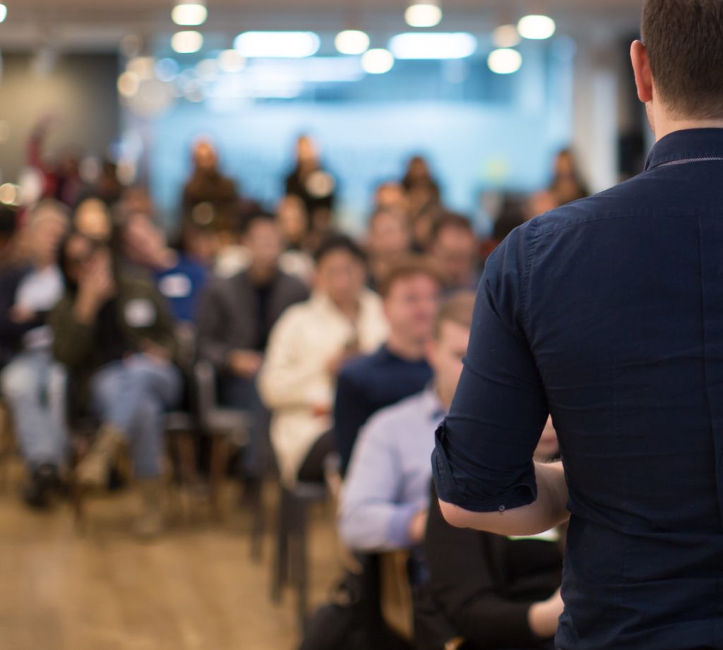 Middle-aged man with his back to camera giving a presentation to a room of 20-30 employees.